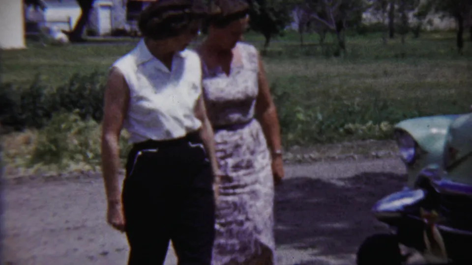 In this 1958 home-movie frame, two women stand by a light green car with lawn and trees behind them.