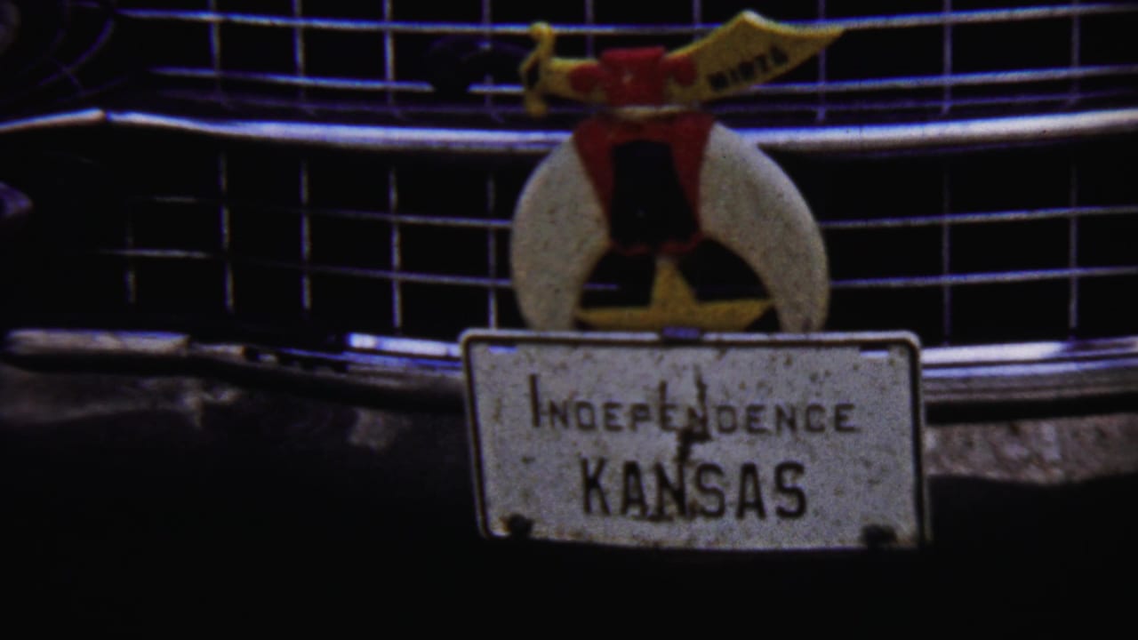 Tight home-movie close-up of the car's chrome grille and readable Independence Kansas plate, with the ornament centered above the plate.