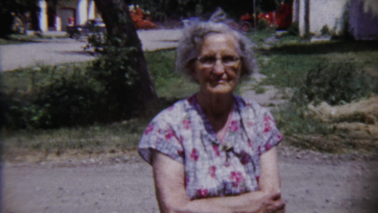 Elderly woman with glasses and gray hair standing outdoors near grass and a path.