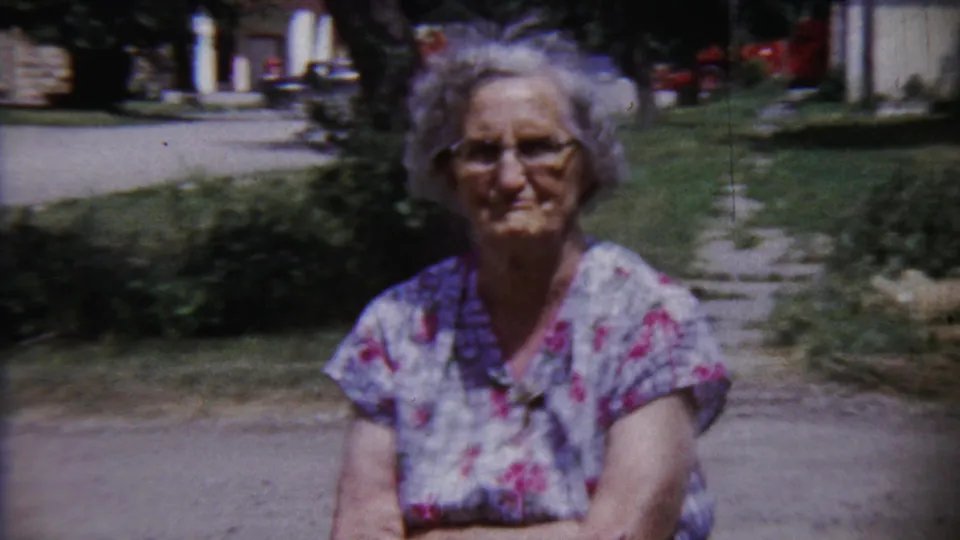 The woman remains posed for the camera in Saint Francis, Kansas, with a path, lawn, shrubbery, and buildings in the background.