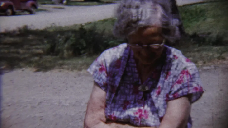 A closer 1958 home-movie frame shows the elderly woman looking down, with her floral blouse, glasses, grass, and path visible.