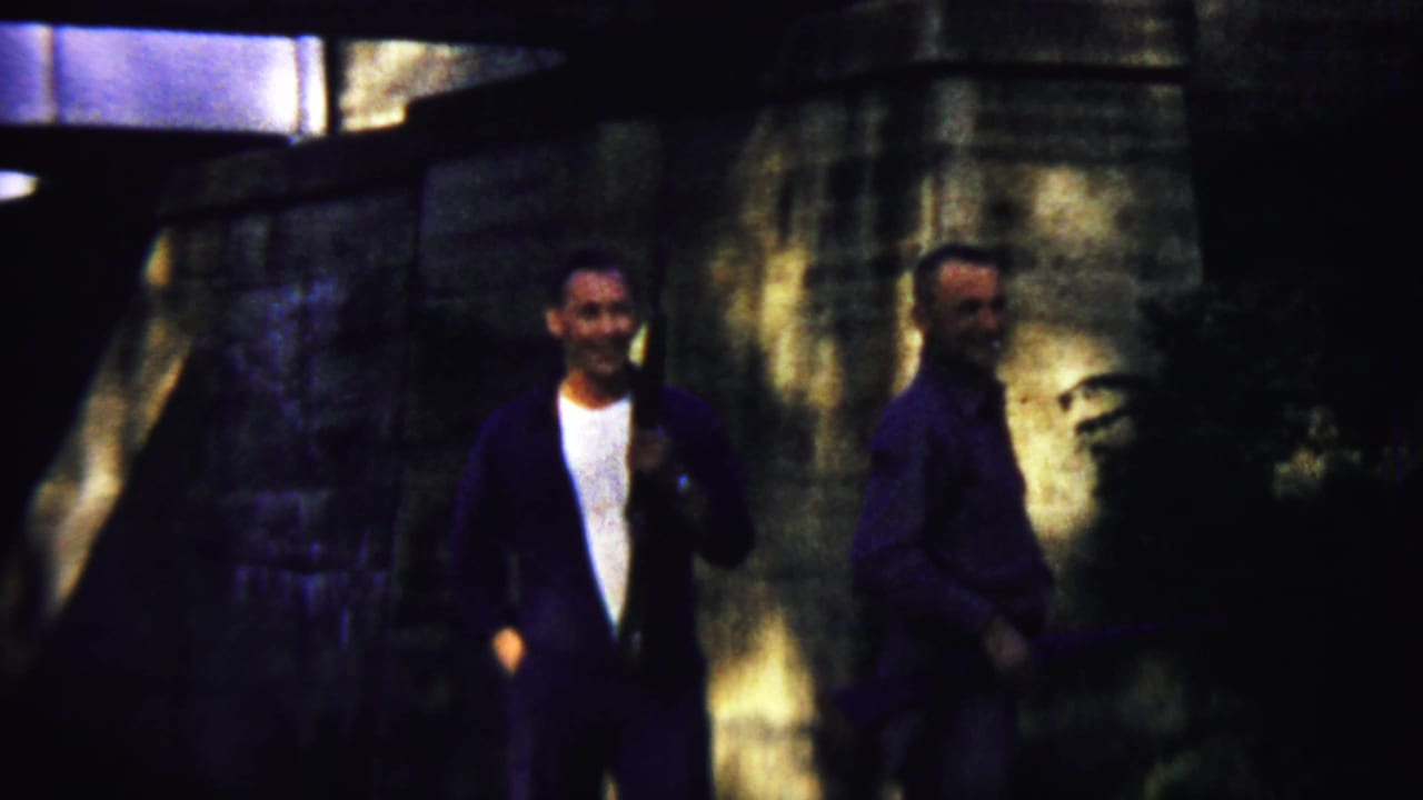 Two adult men stand or walk beneath a stone bridge, one carrying a long gun-like object.