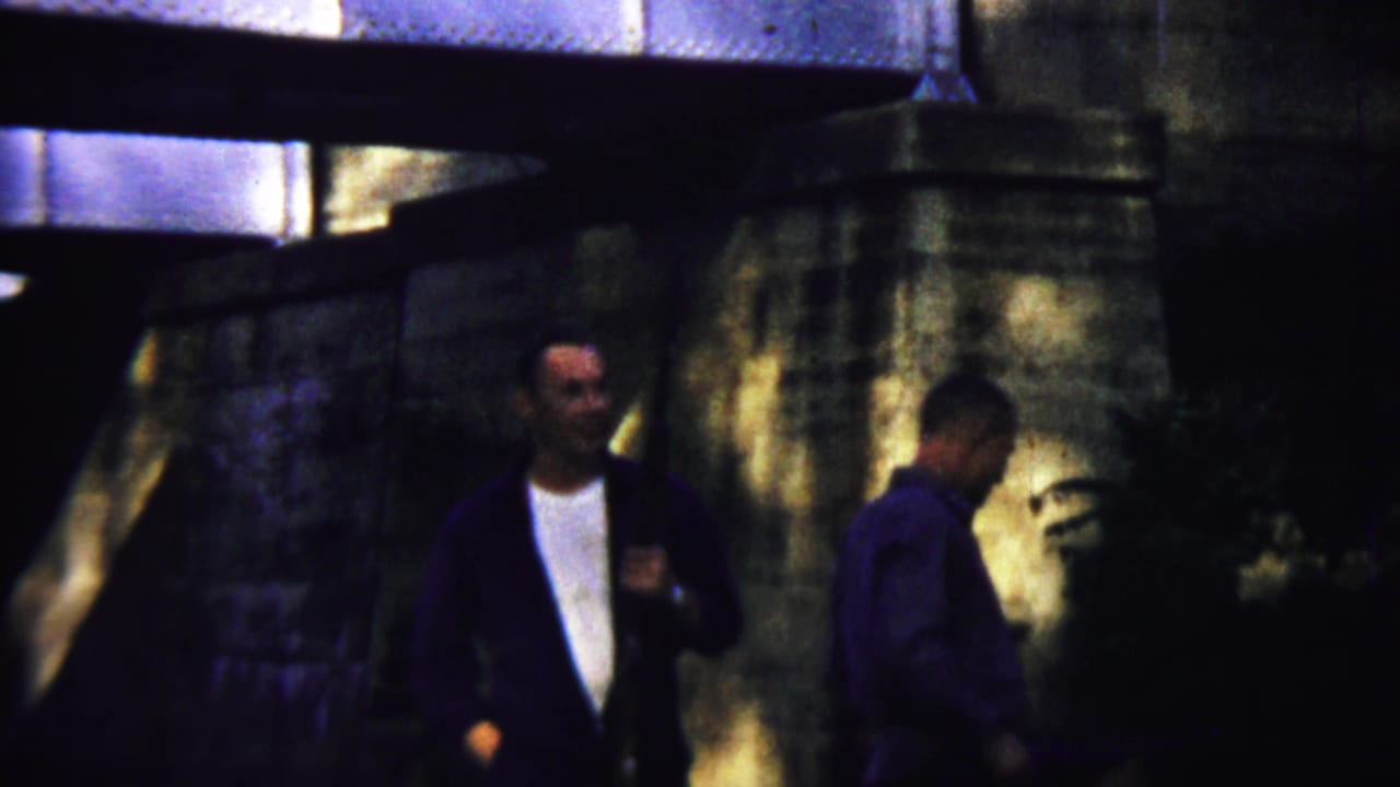 Two men continue walking under the bridge, with one man partly turned away.