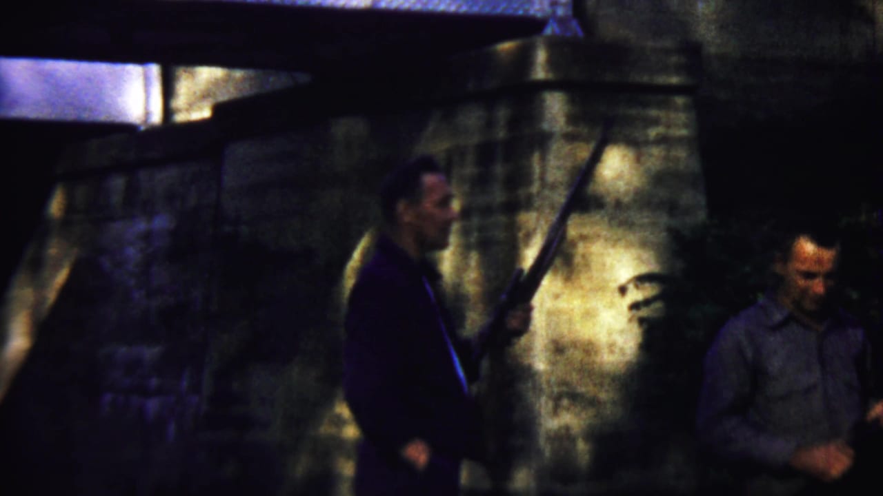 1958 Saint Francis, Kansas home-movie frame showing two adult men near stone masonry, with one man holding a long rifle in low light.
