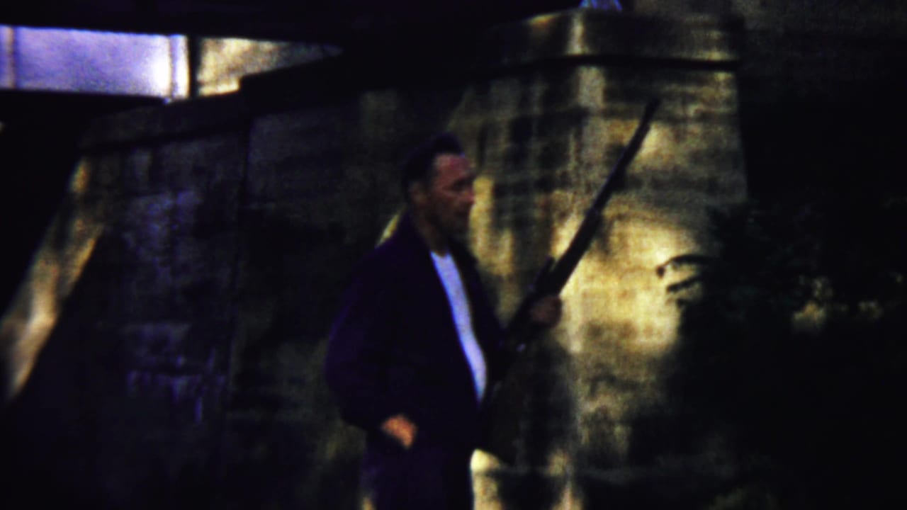 An adult man in a dark jacket holds a long rifle beside stonework.