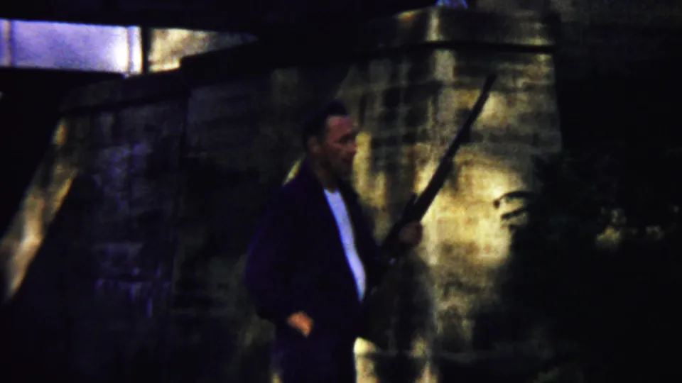 An adult man in a dark jacket and white shirt holds a long rifle near shadowed stone masonry in the 1958 Kansas footage.