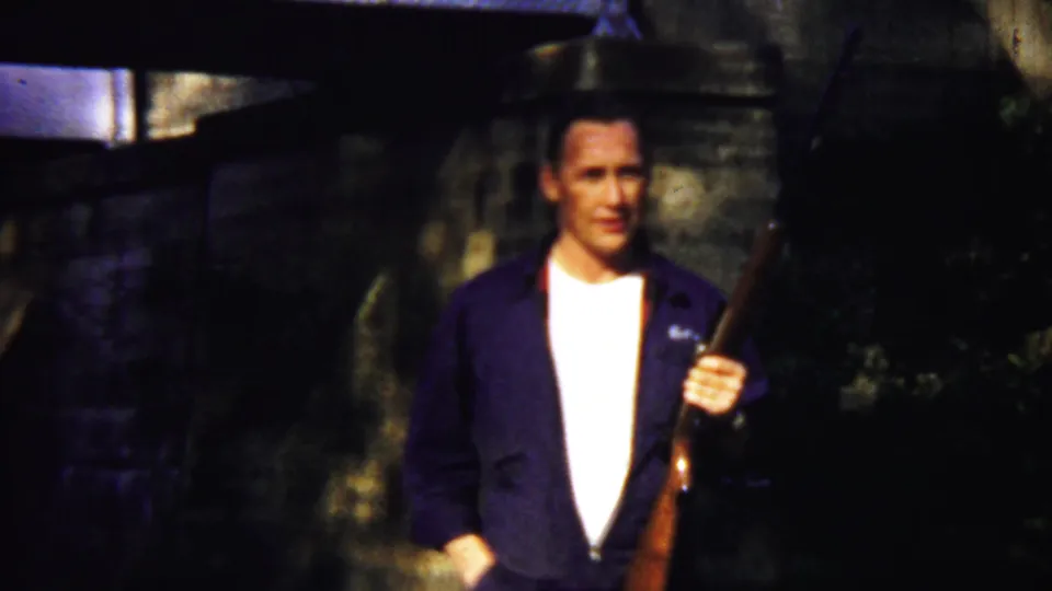 A man in a dark jacket and white shirt faces the camera with a long rifle beside him in the Saint Francis, Kansas home movie.
