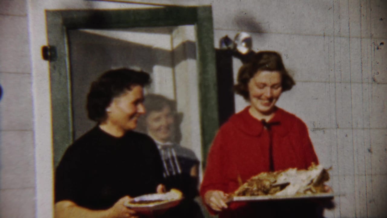 Two women smile while carrying trays of food through an exterior doorway.