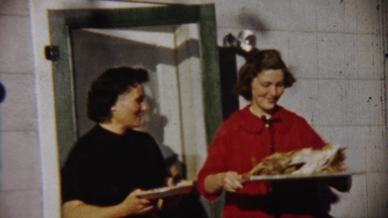 The 1958 Kansas home-movie frame shows two women outside a white-sided house, one holding a plate and one holding a tray with a roasted bird.