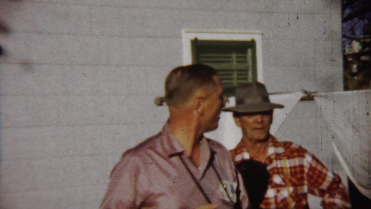 A 1958 Saint Francis, Kansas frame shows two men outdoors, one turned in profile and another in a brimmed hat and plaid shirt near laundry on a line.