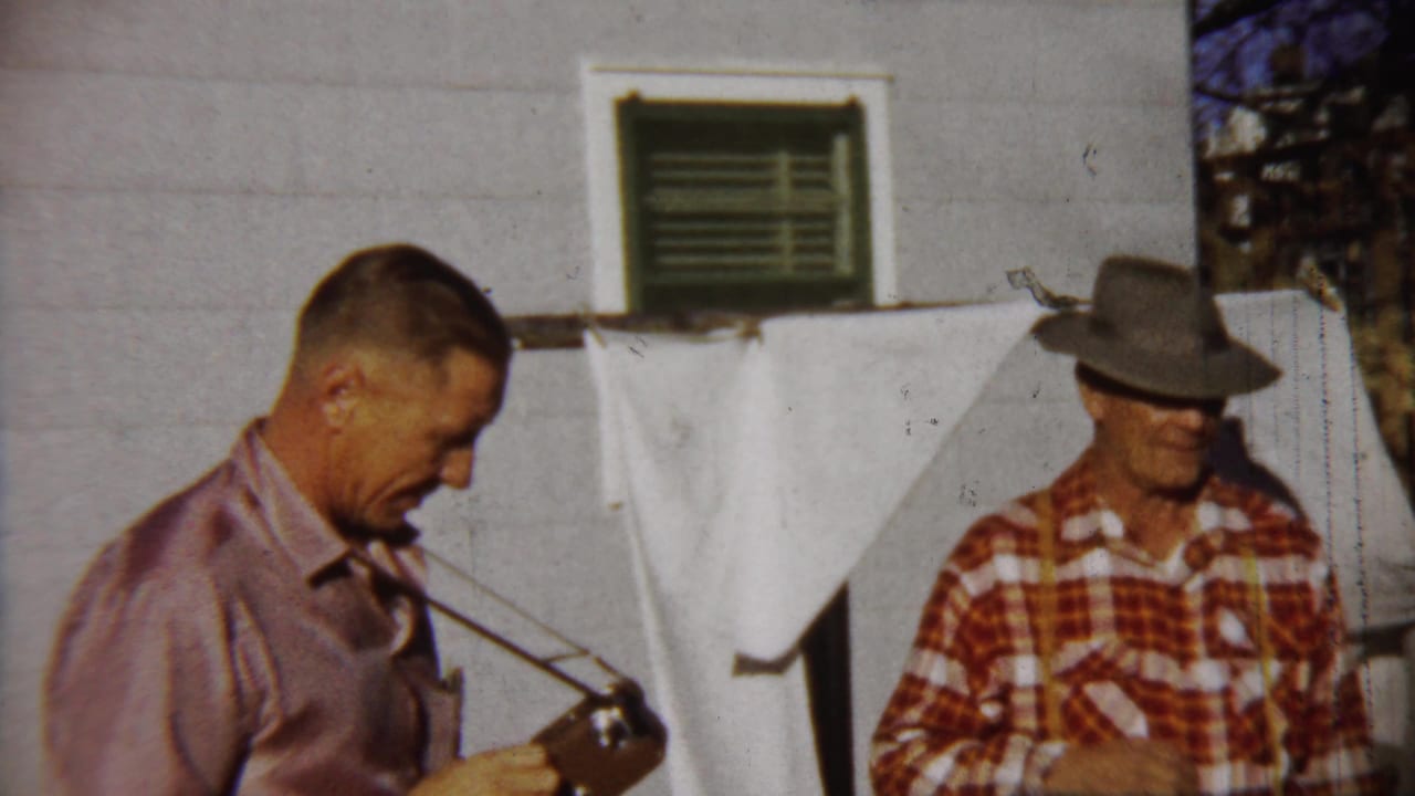 In 1958 Saint Francis, Kansas footage, a man in a pink shirt holds a camera while another man in a fedora stands by a white house.