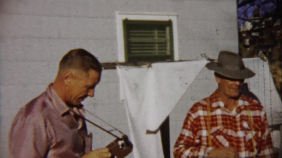 In 1958 Saint Francis, Kansas footage, a man in a pink shirt holds a camera while another man in a fedora stands by a white house.
