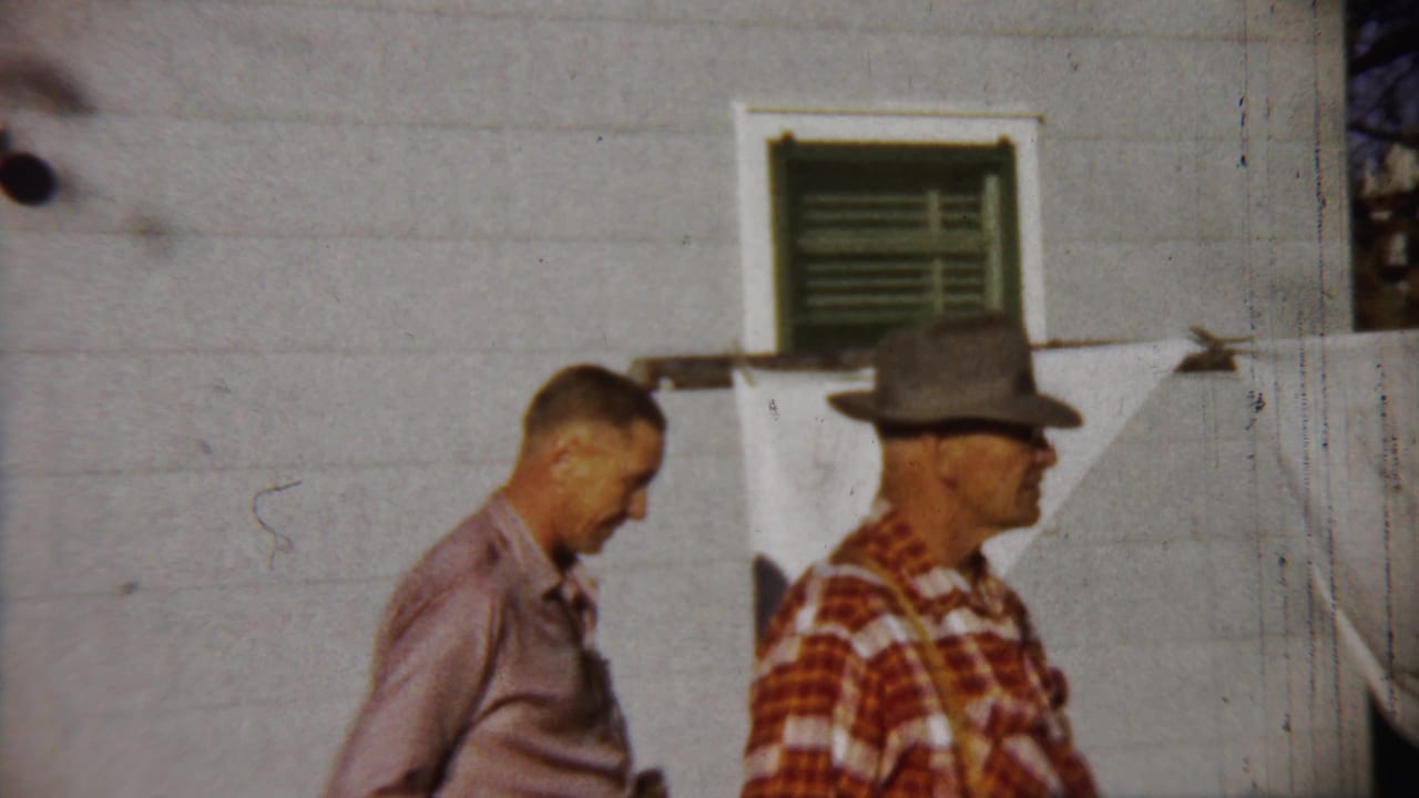 Two adult men in Saint Francis, Kansas pass along a white house exterior, one with a camera and one in a plaid shirt and hat.