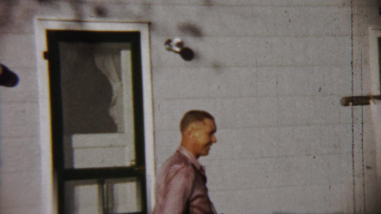A man in a pink shirt appears beside a screen door on a white-sided house in the 1958 Saint Francis, Kansas home movie.