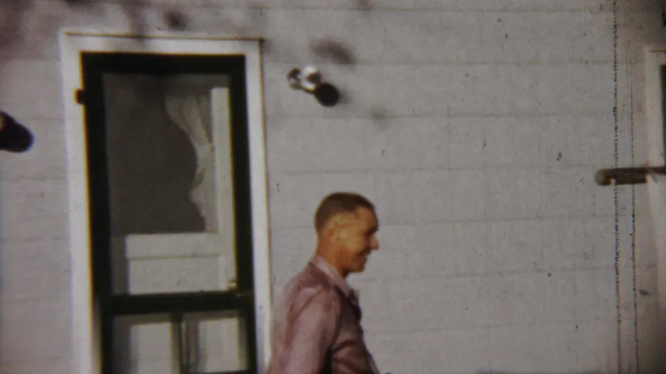 A man in a pink shirt appears beside a screen door on a white-sided house in the 1958 Saint Francis, Kansas home movie.
