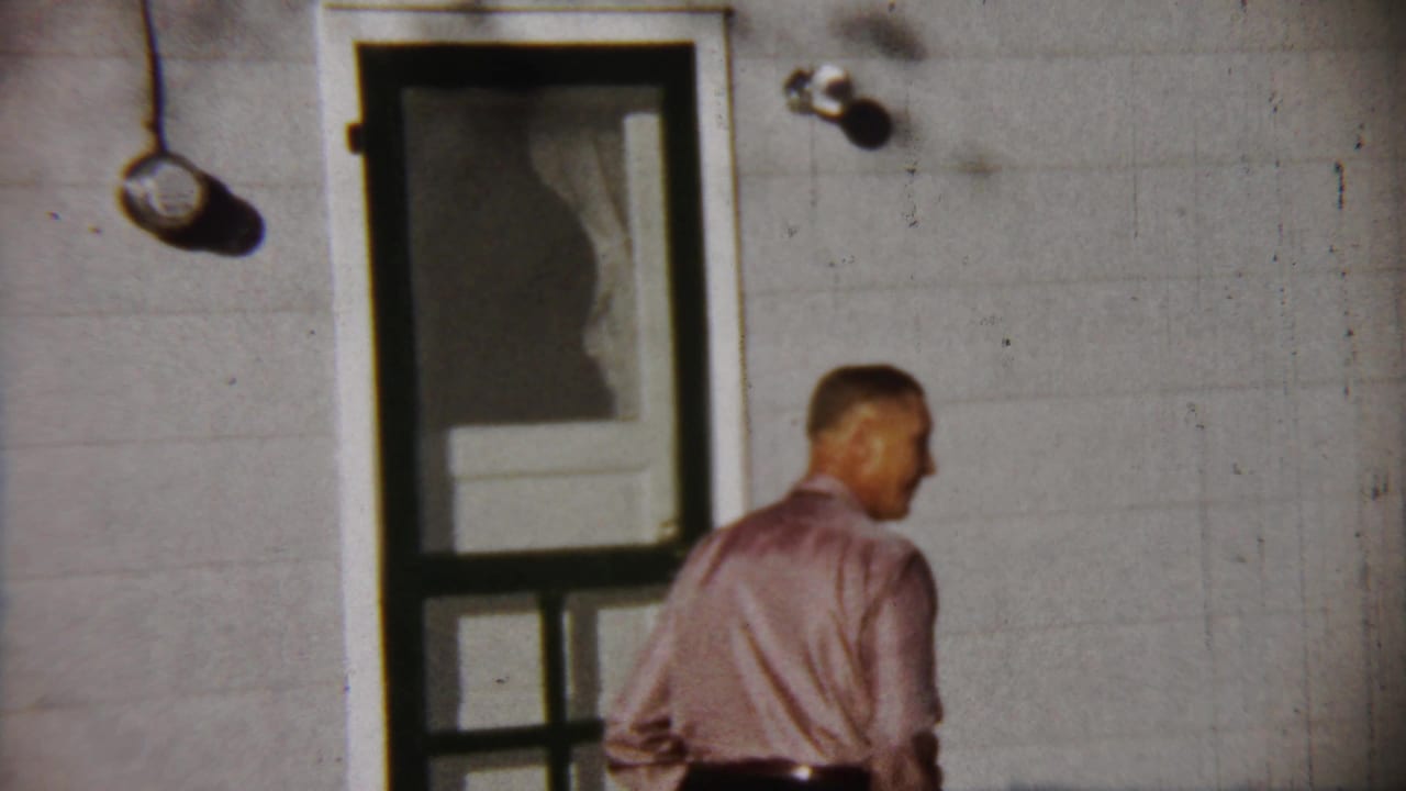 The man in the pink shirt turns near the screen door of a white-sided house in Saint Francis, Kansas home-movie footage.