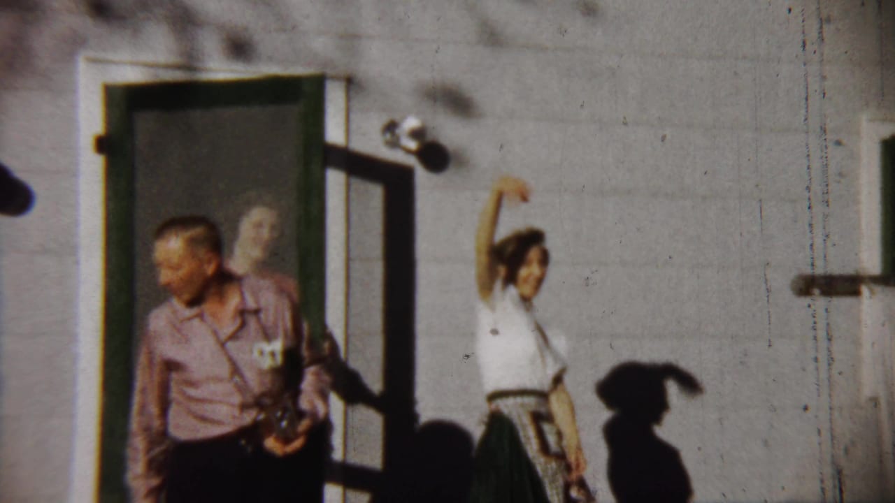 Adults outside a light-colored house as a woman in white raises her arm near an open door.