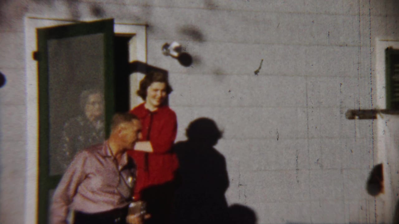 1958 Saint Francis, Kansas frame showing a smiling woman in red at a doorway, with a man in a pink shirt and another person partly visible inside.