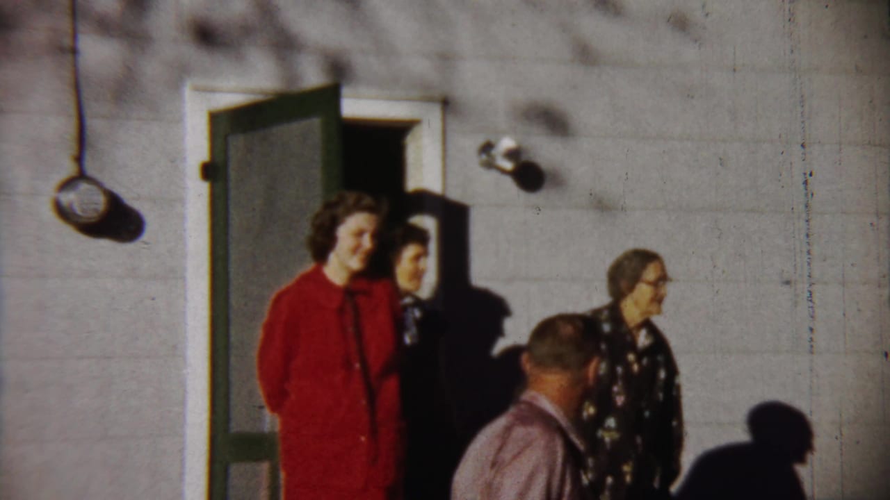 Adults stand near an open green door on a sunlit house exterior.