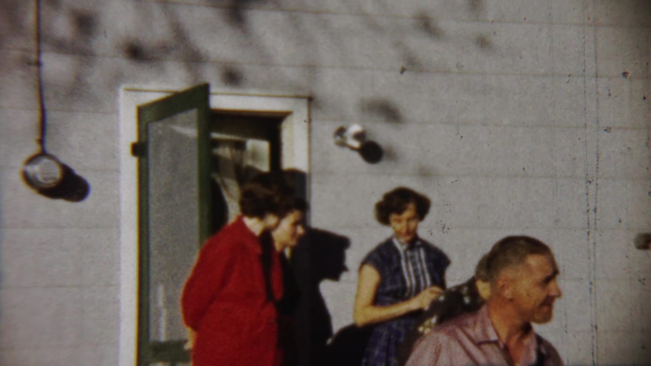 Adults outside a Saint Francis house in 1958, with a woman in red near the open door and a man crossing the foreground.