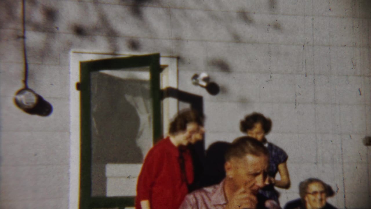 1958 home-movie frame from Saint Francis showing adults outside a house, including a red-dressed woman and a passing man.