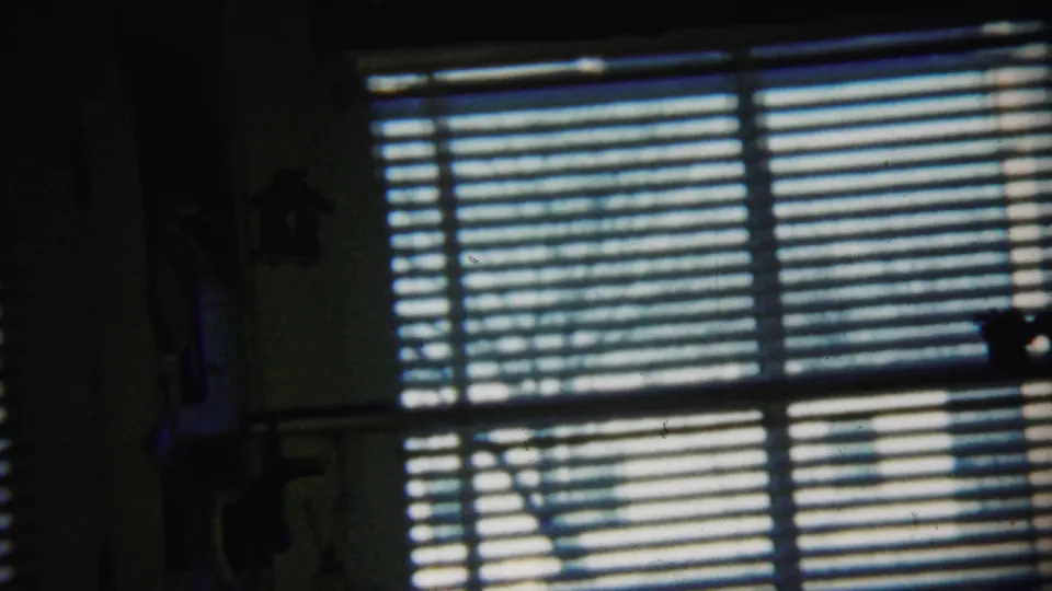 Blue-toned 8mm still of a Saint Francis, Kansas home interior, with venetian blinds and faint silhouettes against daylight.