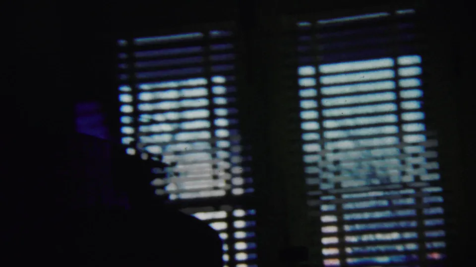 Grainy 1958 interior frame with a bright blinded window and indistinct furniture shapes in shadow.