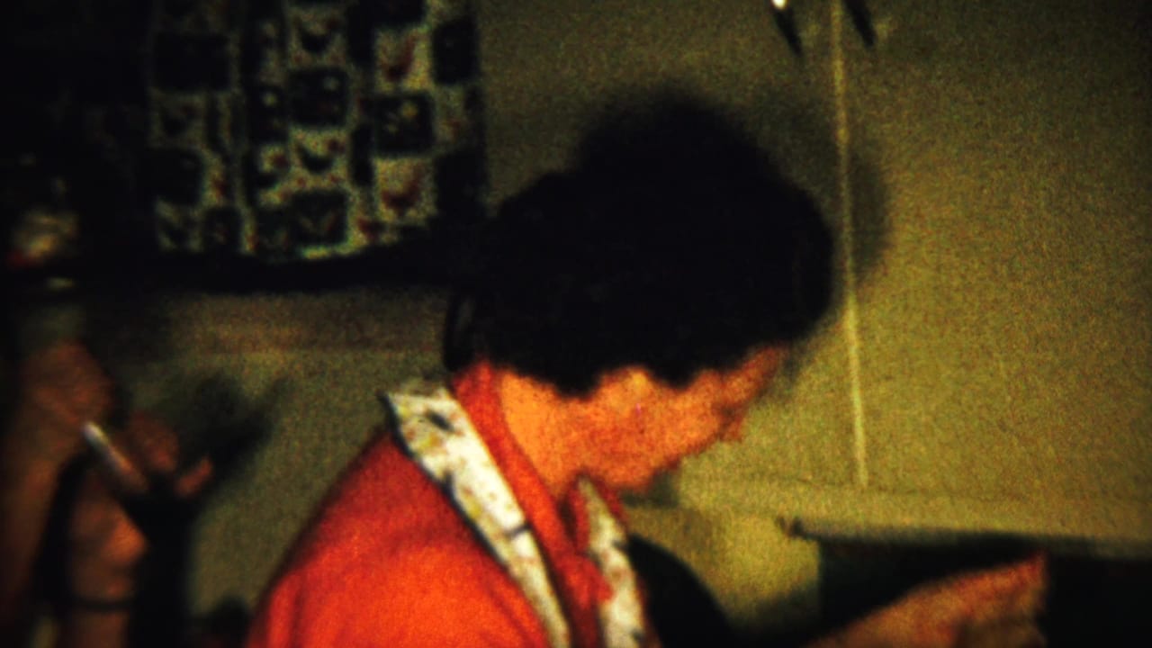 Woman in a red top and apron gestures in a dim kitchen with another person partly visible.