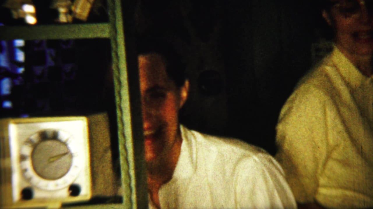 Two adults stand near a small clock radio in a dark kitchen interior.