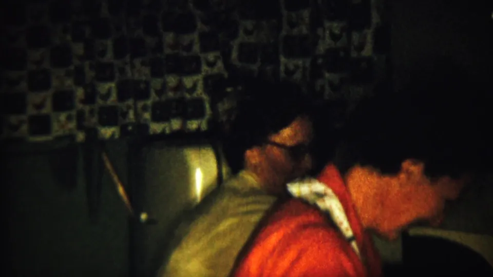 Adults gather in a Saint Francis, Kansas kitchen in 1958, with patterned curtains, a rounded appliance, and a woman in a red top visible.