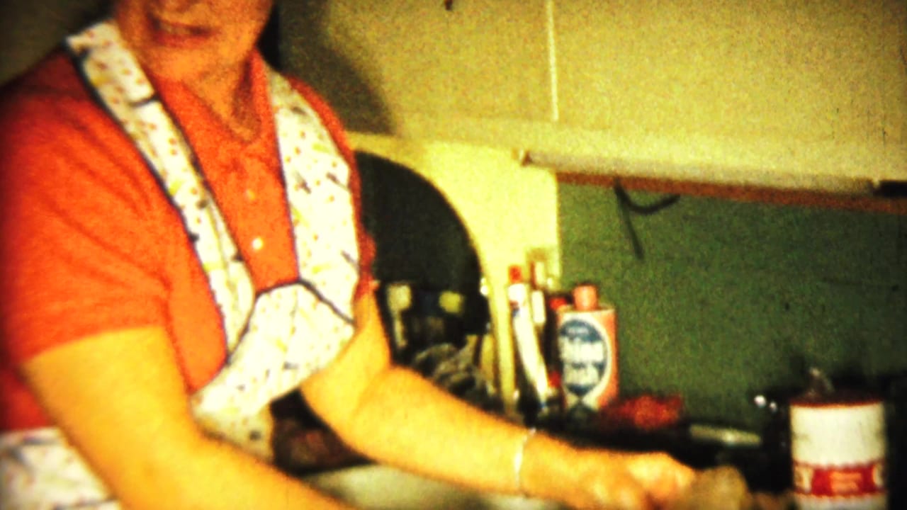 Woman in a red shirt and patterned apron works at a kitchen sink.