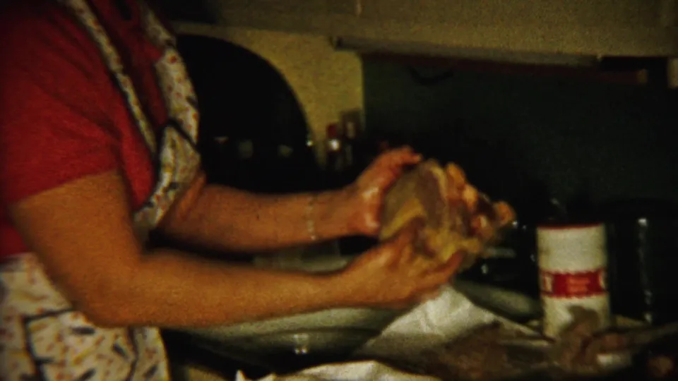 The woman in a patterned apron handles food or dishes over the sink, with kitchen containers and cookware visible nearby.