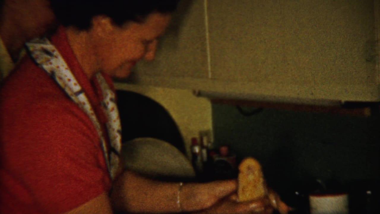 Woman in a red shirt and patterned apron handling food at a dim kitchen counter.