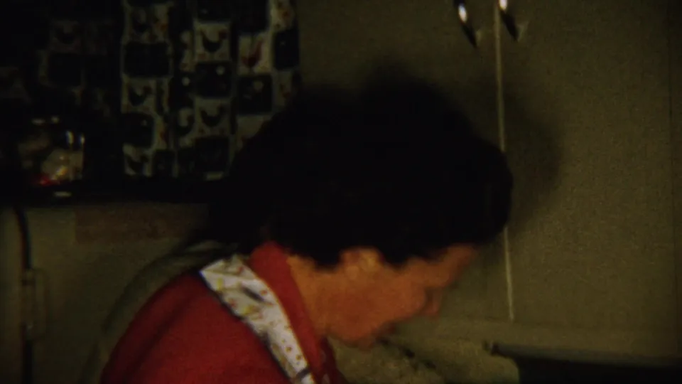 A handheld 8mm frame shows the woman bent over the counter, with bottles and household kitchen items nearby.