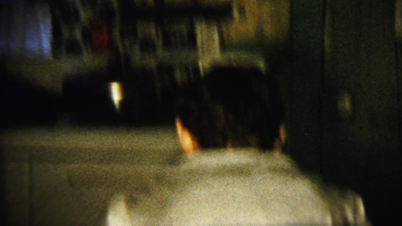 Man in a light shirt bends toward a kitchen counter in a dark interior.