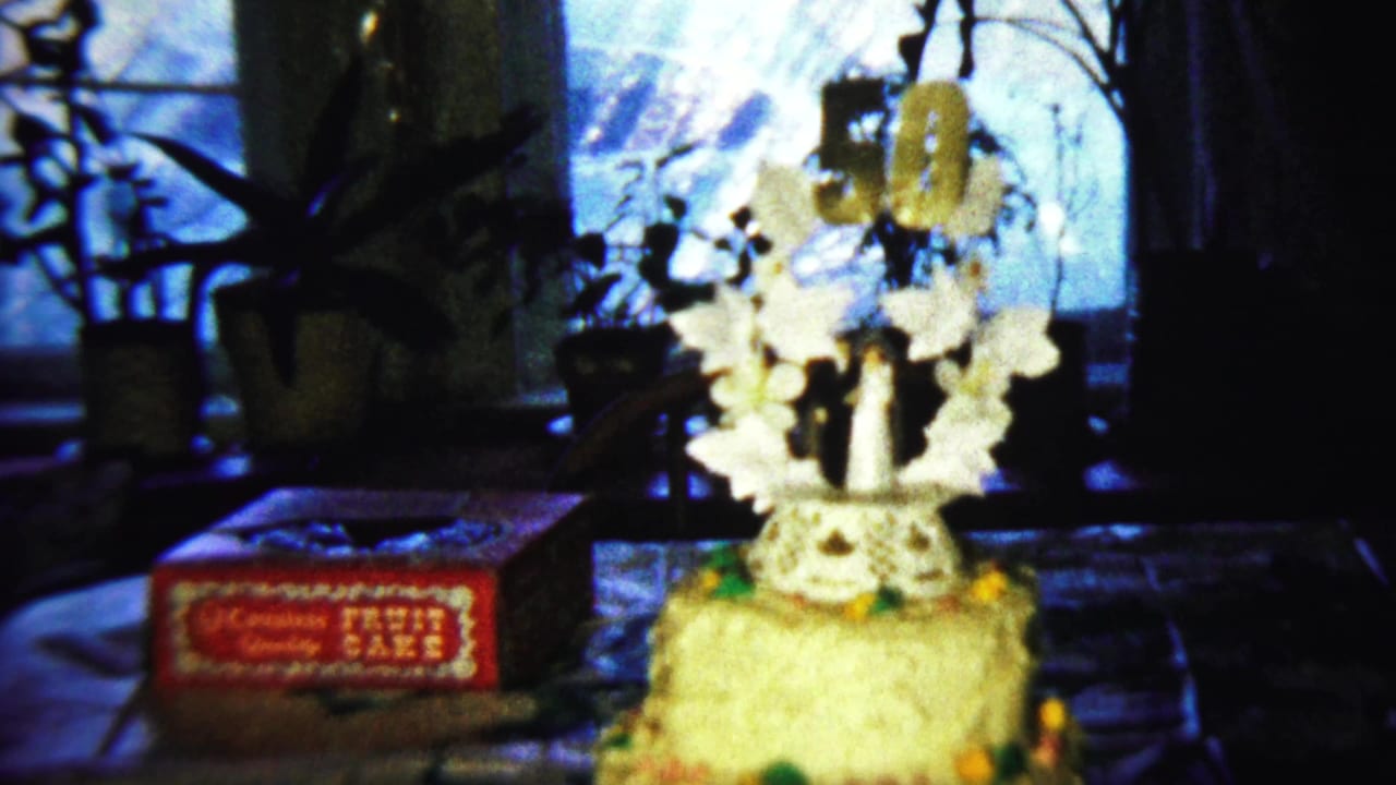 Tiered birthday cake with white floral decorations beside a red fruit cake box on a table.