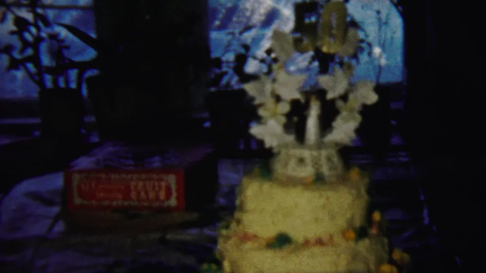 1958 home-movie frame from Saint Francis, Kansas showing the cake from a slightly shifted angle with table, box, and window behind.