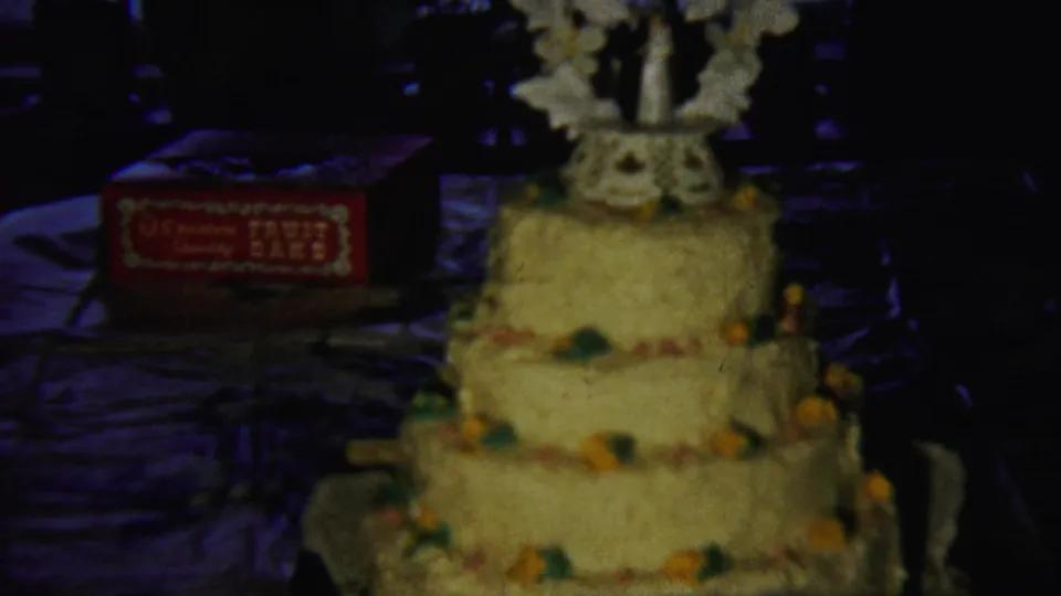 Saint Francis, Kansas 1958 frame showing the decorated yellow birthday cake and nearby red fruit cake box in a dark home interior.