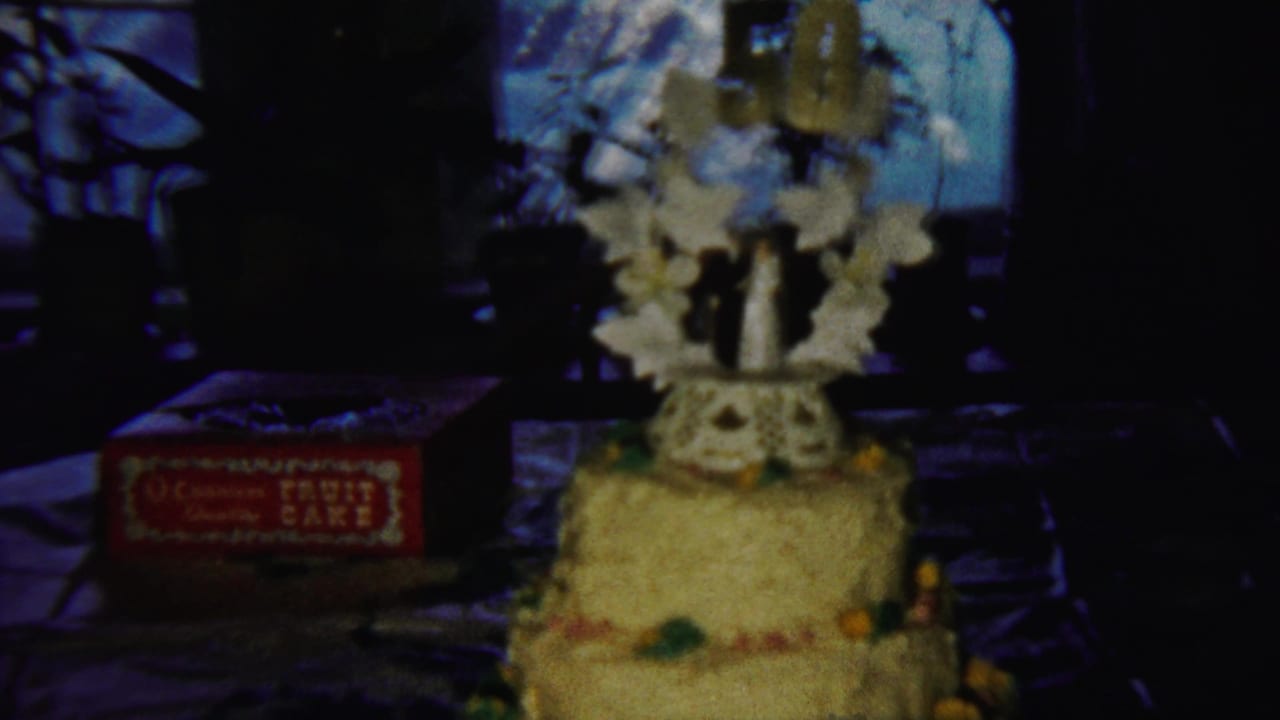 Decorated tiered cake in a dim 1958 Saint Francis, Kansas home-movie frame, with a red boxed fruit cake beside it.