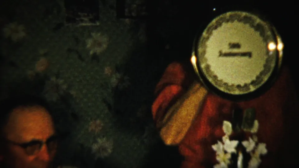 Final home-movie frame shows the anniversary plate clearly raised beside a seated man in glasses in a private interior.