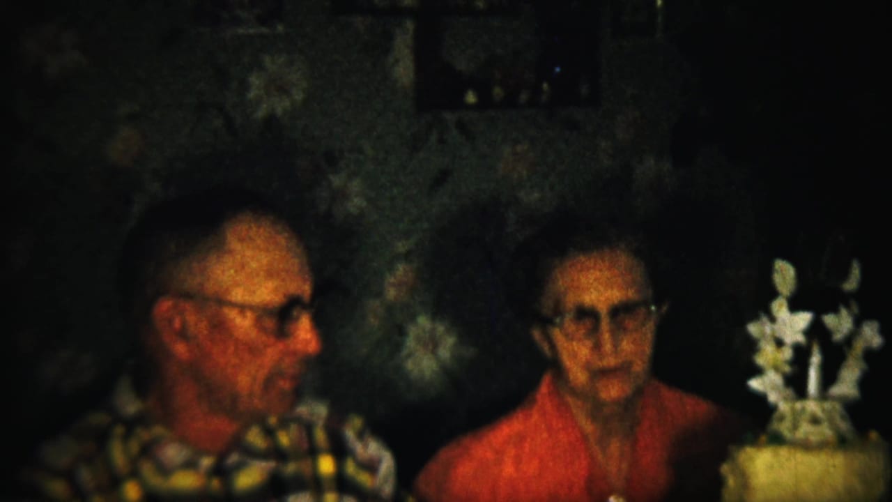 Man in glasses sits beside a woman in a dim room with a white floral decoration nearby.