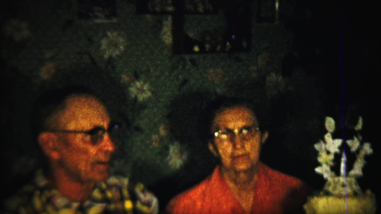 Older man in glasses faces sideways while a woman in red sits beside a white floral decoration.