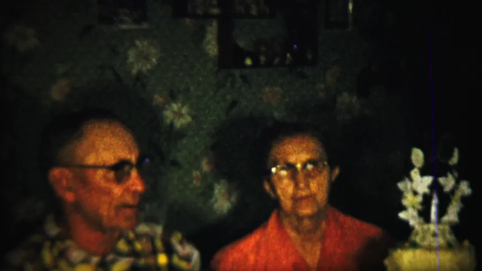 1958 Saint Francis, Kansas home-movie frame with an older man in glasses in profile and a woman in a red top seated nearby indoors.