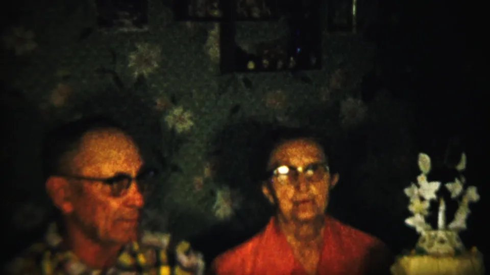 1958 Saint Francis, Kansas 8mm frame showing the seated adults more frontally, with glasses catching light and a white floral decoration at right.