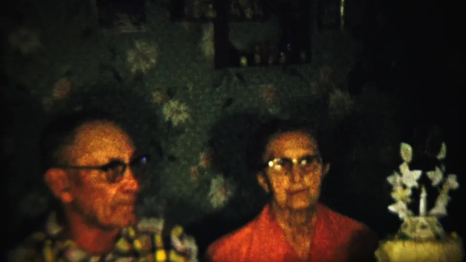 1958 Saint Francis, Kansas home-movie frame of two adults seated indoors, with the man turned slightly and the woman looking forward beside a white decoration.