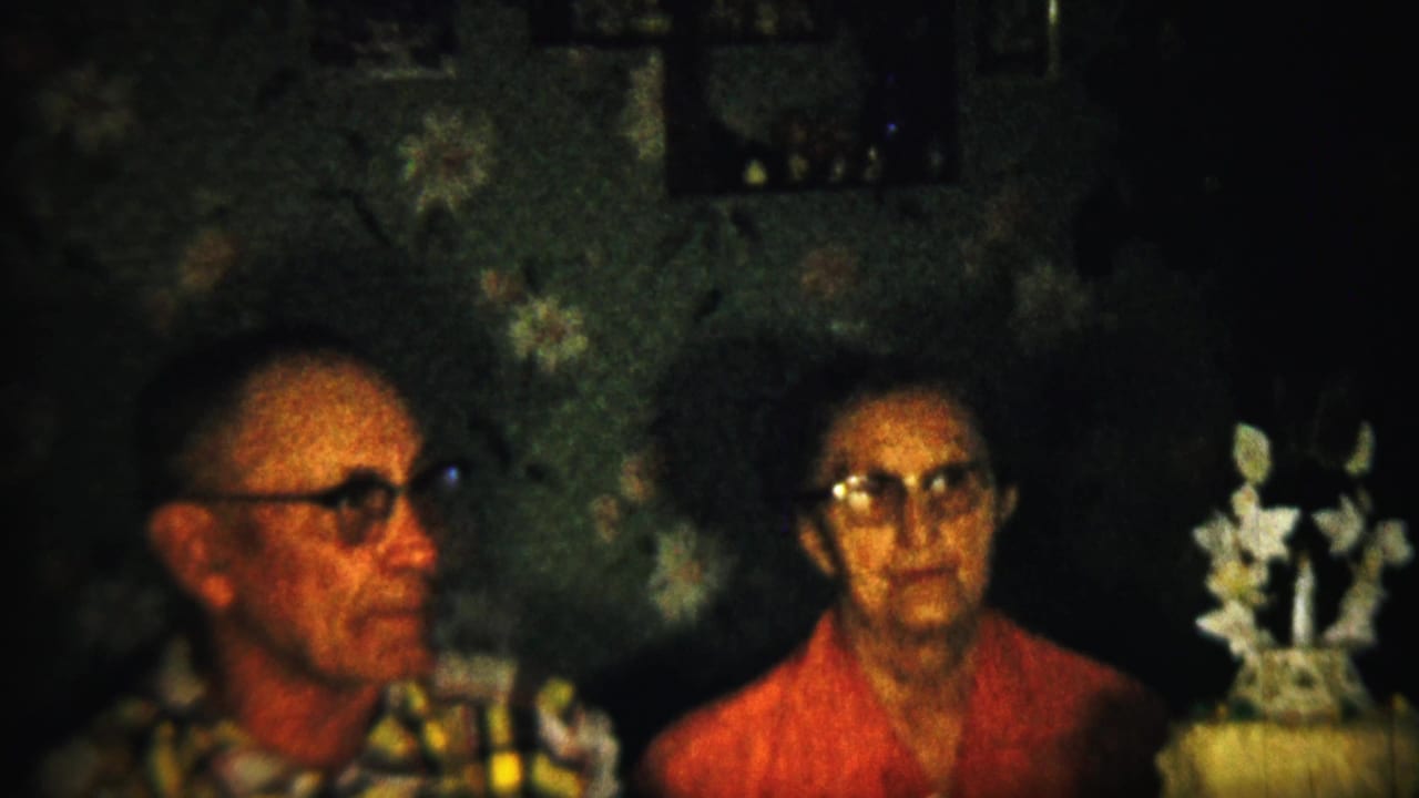 In this 1958 Saint Francis, Kansas home-movie frame, an elderly man in glasses and an elderly woman in a red top sit indoors beside a decorated cake with white floral ornaments.