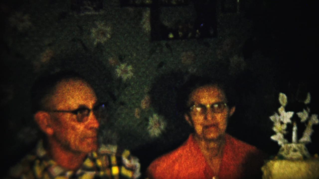 A soft 8mm frame shows the older couple seated against floral wallpaper, the woman facing forward and the decorated cake with white ornaments positioned beside her.