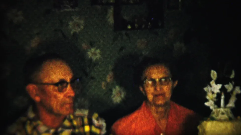 A soft 8mm frame shows the older couple seated against floral wallpaper, the woman facing forward and the decorated cake with white ornaments positioned beside her.