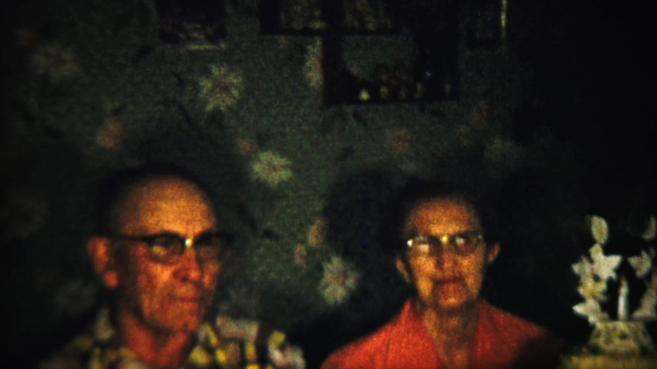 The final frame holds on the elderly couple in the dim interior, with the man in profile, the woman facing forward, and the decorated cake still visible on the right.