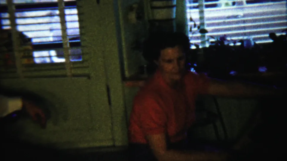 In Saint Francis, Kansas home-movie footage from 1958, a woman in a red blouse sits in a dim private interior near window blinds.